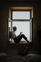 MAn reading a book while sitting in a window.